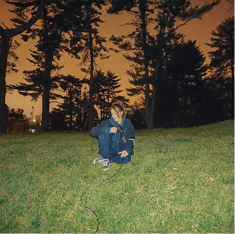 Album cover: a person crouched in a grassy field at dusk, holding a microphone cable